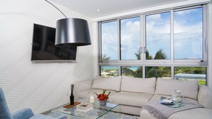 A modern living room with a sofa, a glass coffee table, a blue chair, and a large window offering a view of the outdoors. - ?Blue Haven? | Oceanfront + Beach View | Pool, Gym (Eagle Beach)