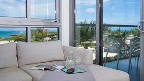 A modern living room with a large window overlooking a beach and ocean, a sofa with a magazine, a pitcher of water, and a glass of white wine. - ?Blue Haven? | Oceanfront + Beach View | Pool, Gym (Eagle Beach)