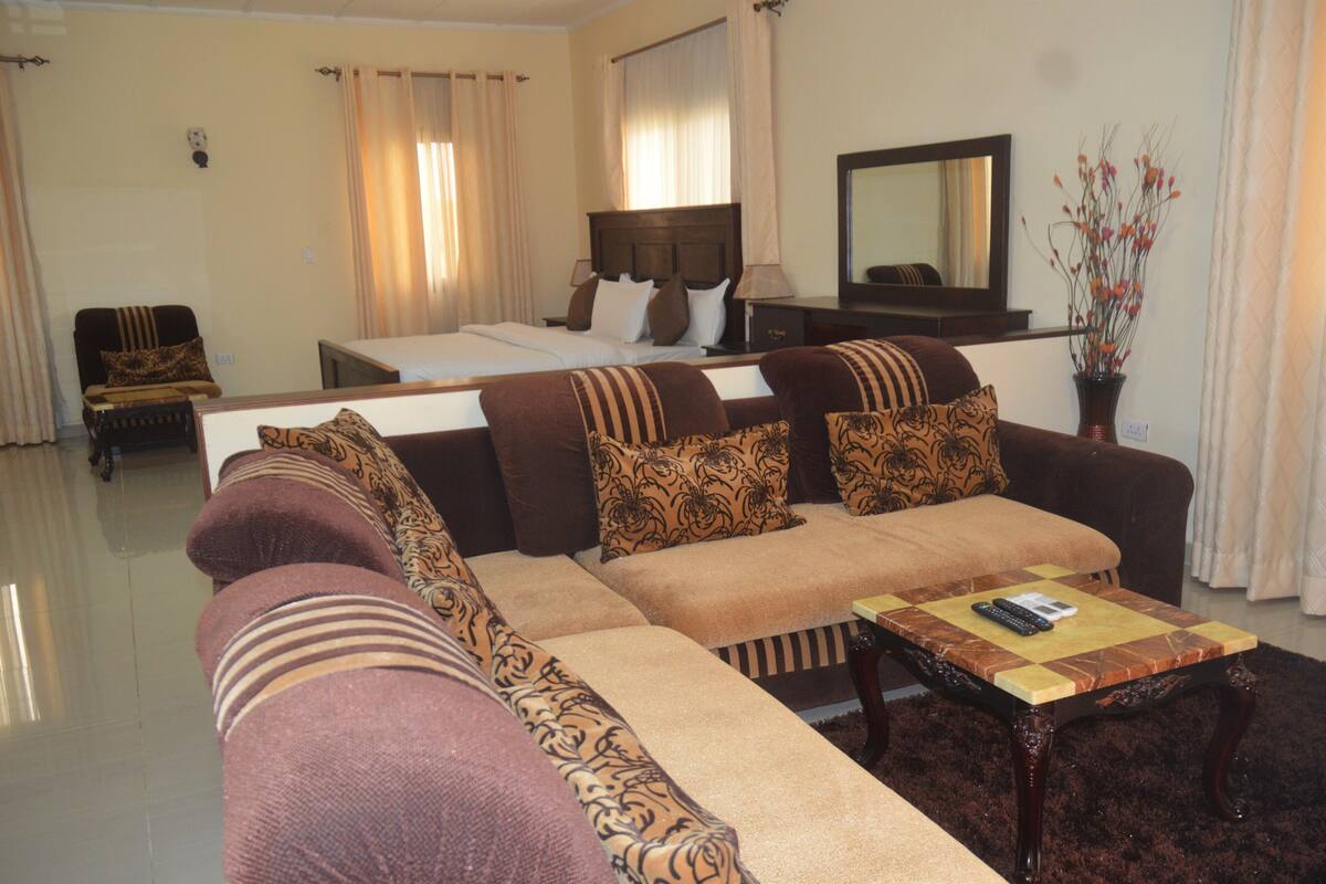 A hotel room with a large sofa, a bed, a small table with a remote control, and a mirror on the wall.