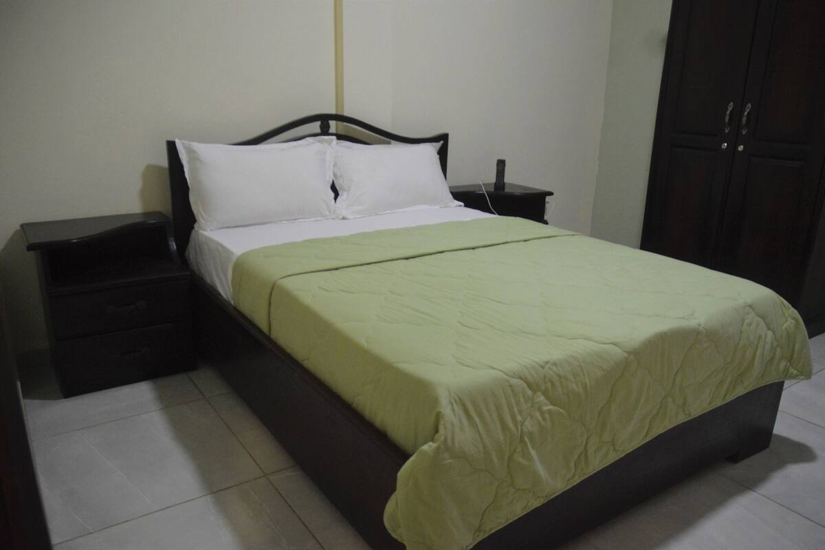 A single bed with a green bedspread and white pillows, a bedside table, and a dark wooden wardrobe.