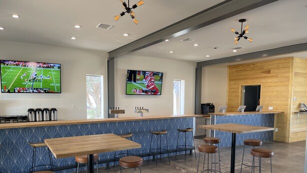 A modern bar area with wooden tables and stools, a counter with a digital screen, and a kitchen area with wooden cabinetry. - The Armadillo Hotel (Odessa)