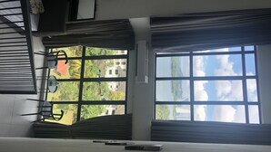 Ett rum med ett stort fönster som ger utsikt över byggnader och grönområden. Det finns en säng med en sänggavel, ett sängbord och en stol.