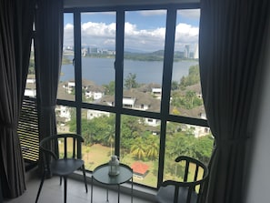 A room with a large window offering a view of a lake, buildings, and greenery. There are two chairs and a small table with a lantern. - White Ivy Sawtelle Suites Cyberjaya (Cyberjaya)