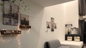 Ett modernt vardagsrum med en grå soffa, ett litet runt bord och en säng med matchande kuddar. På väggarna hänger konstverk som föreställer berömda landmärken, och en ljusslinga hänger längs väggen.