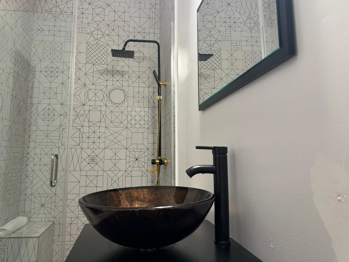 Une salle de bain avec un mur de douche orné d’un motif géométrique, un lavabo noir et un miroir.