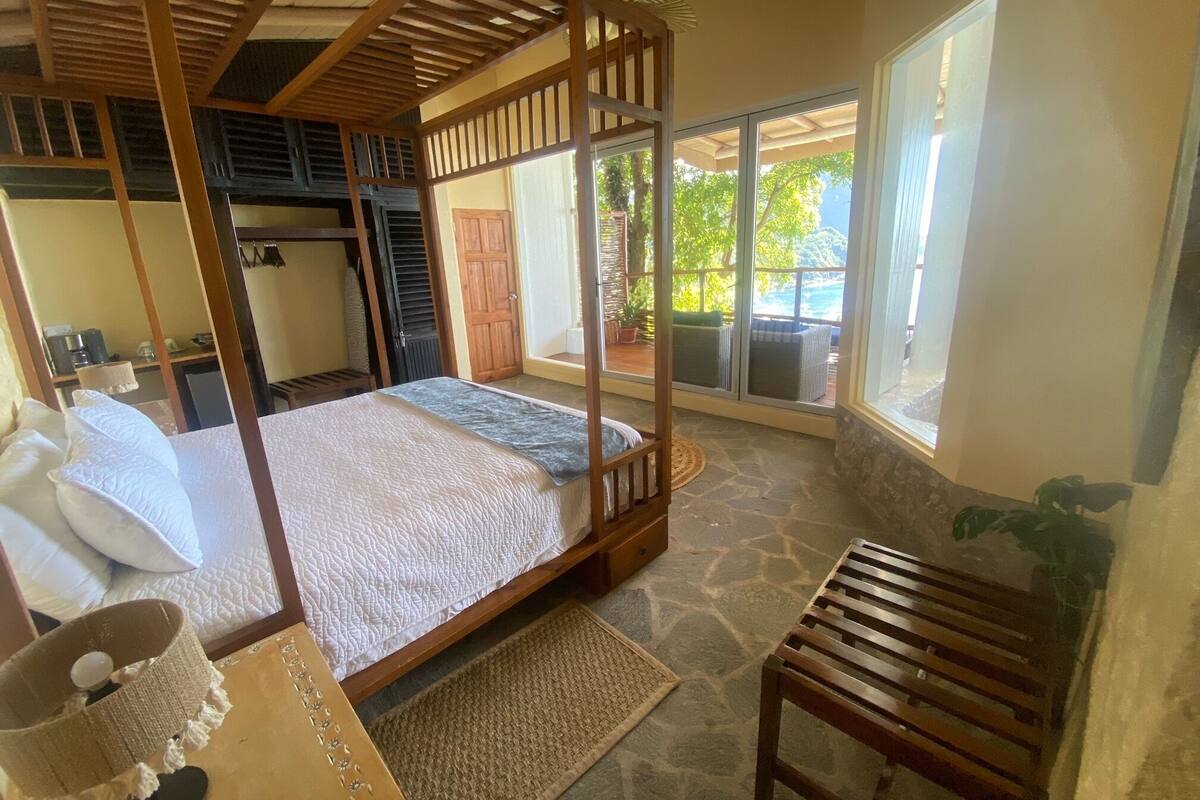 A bedroom with a four-poster bed, a wooden bench, a stone floor, and a view of the outdoors through large windows.