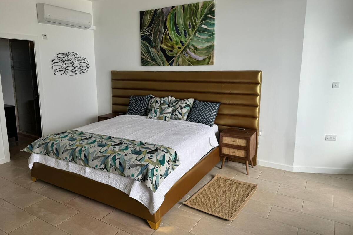 A bedroom with a bed, a headboard, a nightstand, a wall-mounted air conditioner, a wall art piece, and a door.