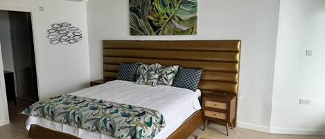 A bedroom with a bed, a headboard, a nightstand, a wall-mounted air conditioner, a wall art piece, and a door.