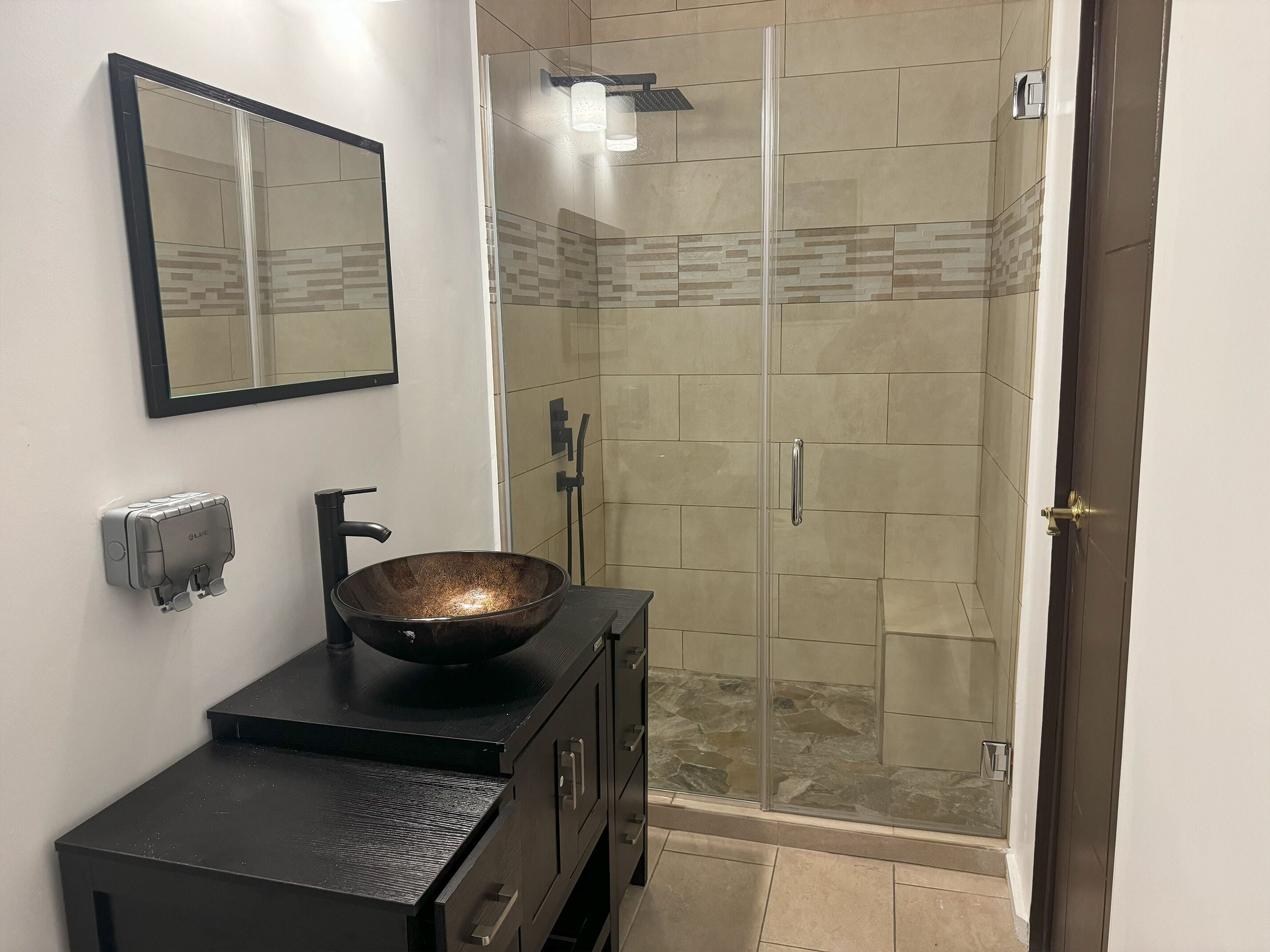 Une salle de bain avec une douche à parois de verre, un lavabo noir doté d’une cuve en cuivre, un miroir et un distributeur de savon.