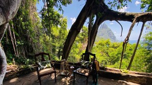 Un patio de piedra con dos sillas de madera y una mesa, rodeado de vegetación exuberante y vistas a un paisaje montañoso.