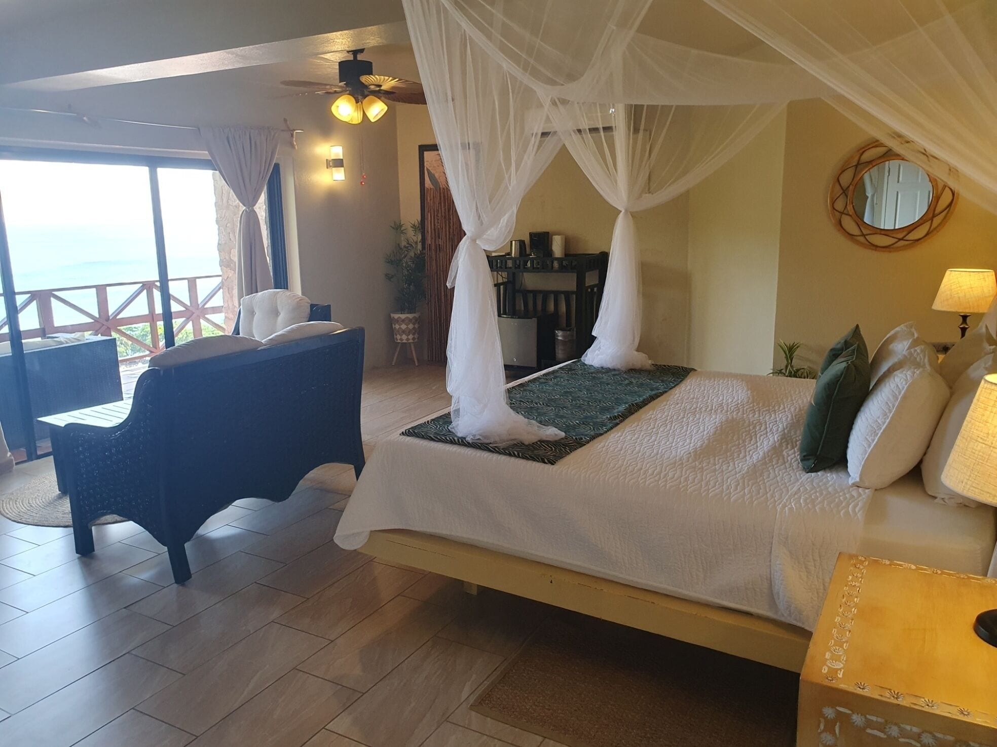 A bedroom with a bed, a chair, a bedside table, a lamp, and a view of the ocean.
