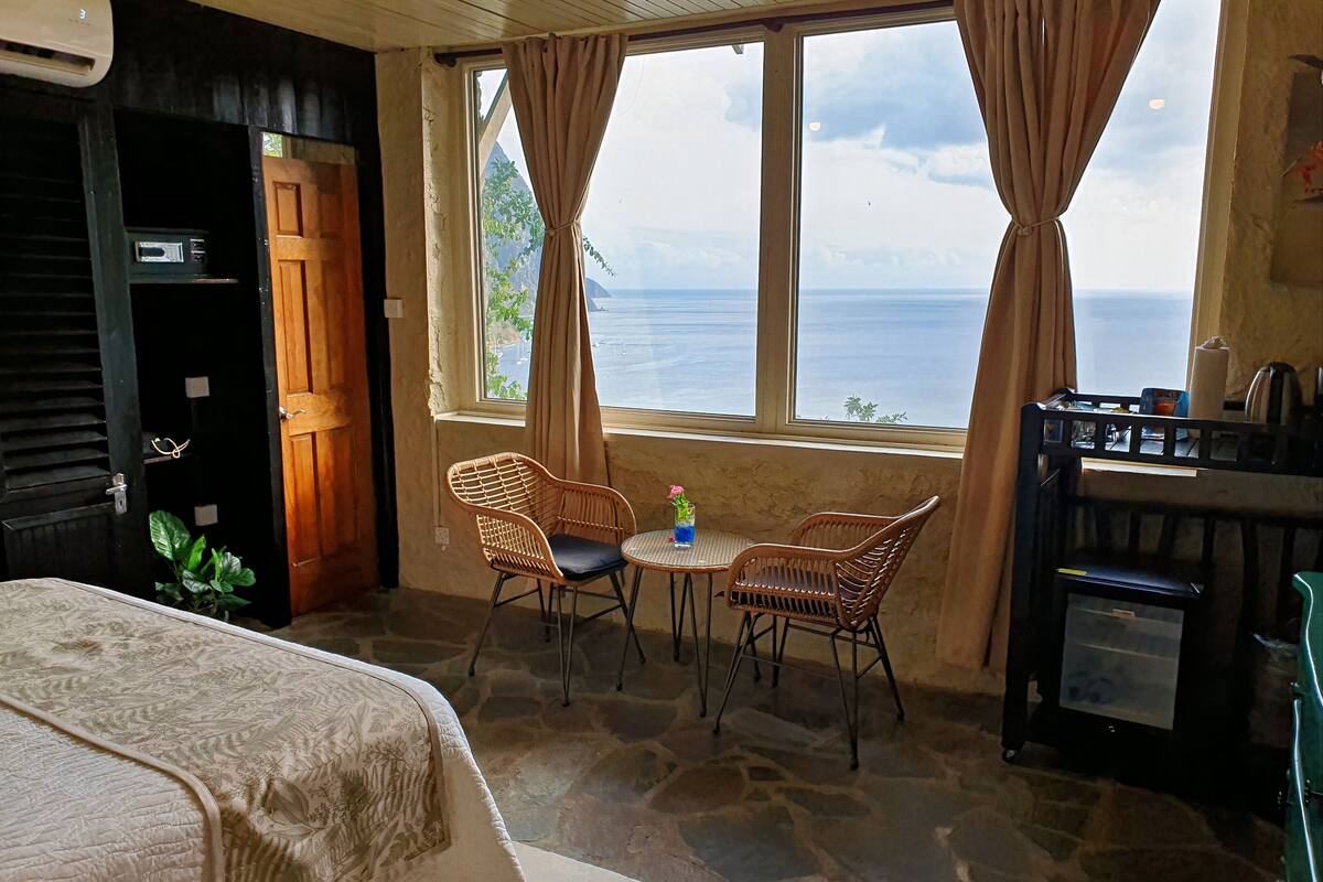 A room with a bed, a table with two wicker chairs, a view of the sea, and a small kitchenette.