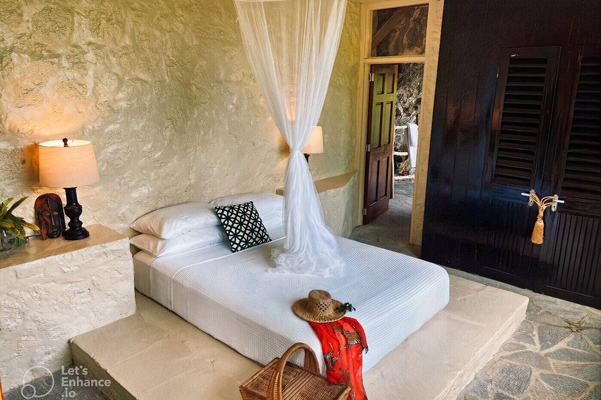 A bed with a canopy, a bedside lamp, a patterned pillow, and a straw hat on the floor.