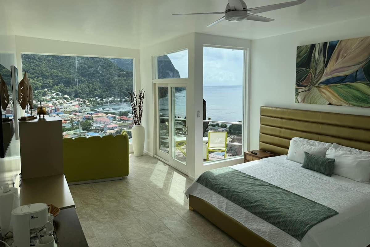 A spacious bedroom with a large bed, a ceiling fan, and a view of a coastal town through large windows.