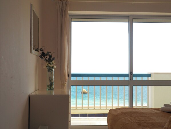 Une chambre avec un lit, une petite table sur laquelle est posé un vase, une fenêtre donnant sur la mer et une balustrade de balcon.