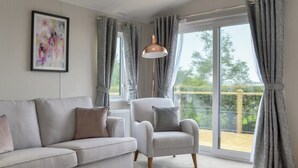 A living room with a grey sofa, a grey armchair, and a floor lamp. There is a window with grey curtains and a view of greenery outside. - 2 bedroom accommodation in Calthwaite, near Penrith (Calthwaite, near Penrith)