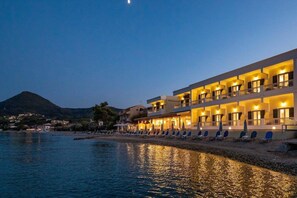 A hotel with multiple balconies overlooking a beach, a pool area with lounge chairs, and a mountain in the background. - Rossi S Studios and Apartments (Corfu)
