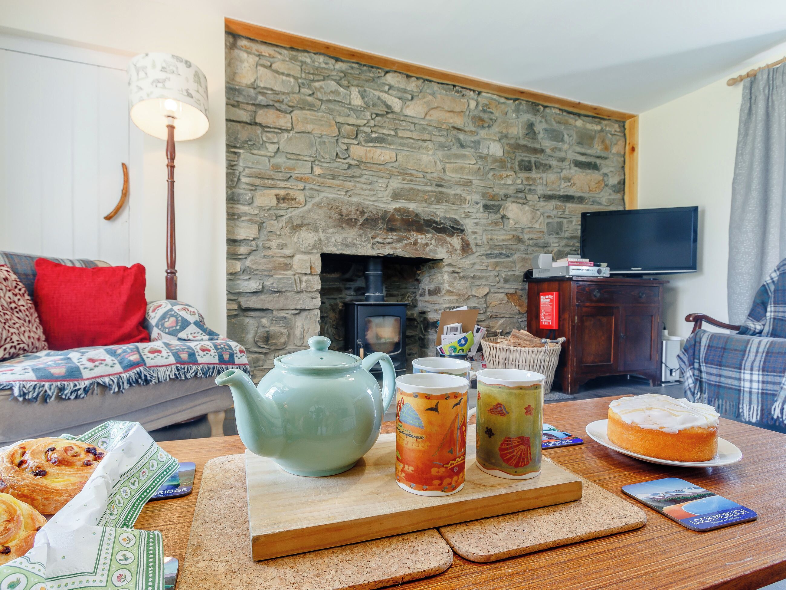 A cozy living room with a stone fireplace, a sofa with patterned cushions, a wooden coffee table with pastries and a teapot, and a television on a wooden cabinet.