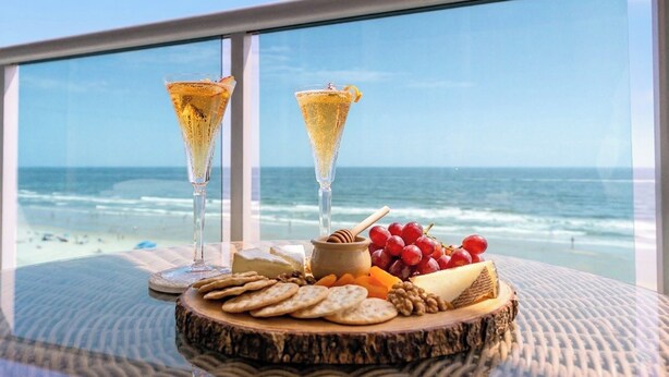 A table with a cheese platter, crackers, grapes, and two glasses of champagne with a view of the ocean.