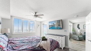 A bedroom with a bed, a ceiling fan, a painting, and a view of the outdoors.