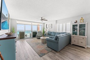 A spacious living area with a large window, a ceiling fan, a blue sofa, a wooden coffee table, and a cabinet with glass doors.