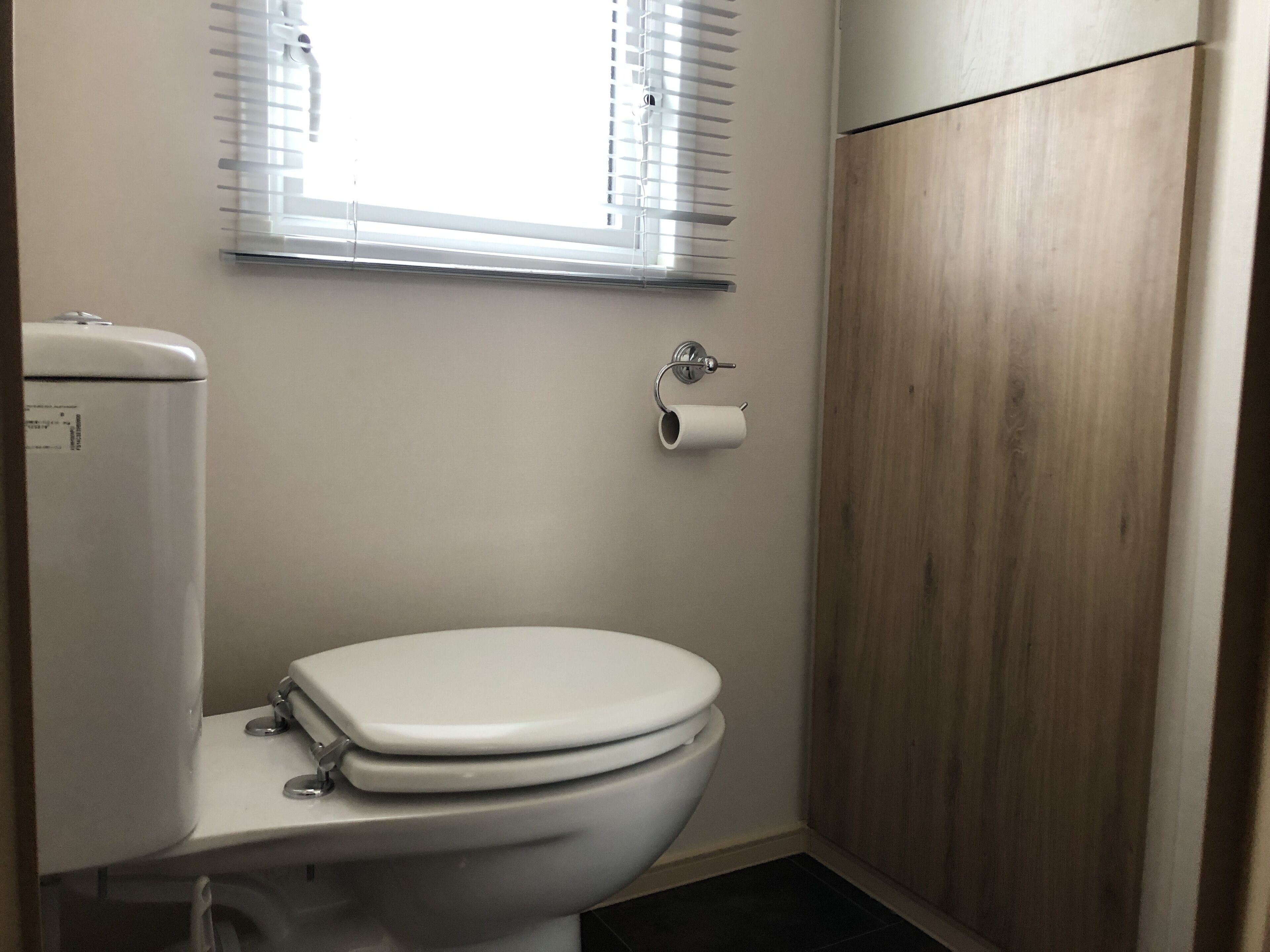 A modern bathroom with a white toilet, a toilet paper holder, and a window with blinds.