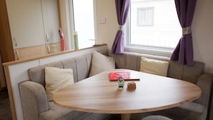 A cozy seating area with a round wooden table, cushioned sofas, and a window with purple curtains. - Excellent Caravan 4 Birth Sleeps 6 Tranquil Site (Colchester)