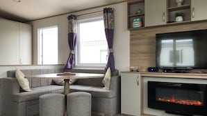 A cozy living area with a sofa, a round table, a television, and a fireplace. - Excellent Caravan 4 Birth Sleeps 6 Tranquil Site (Colchester)