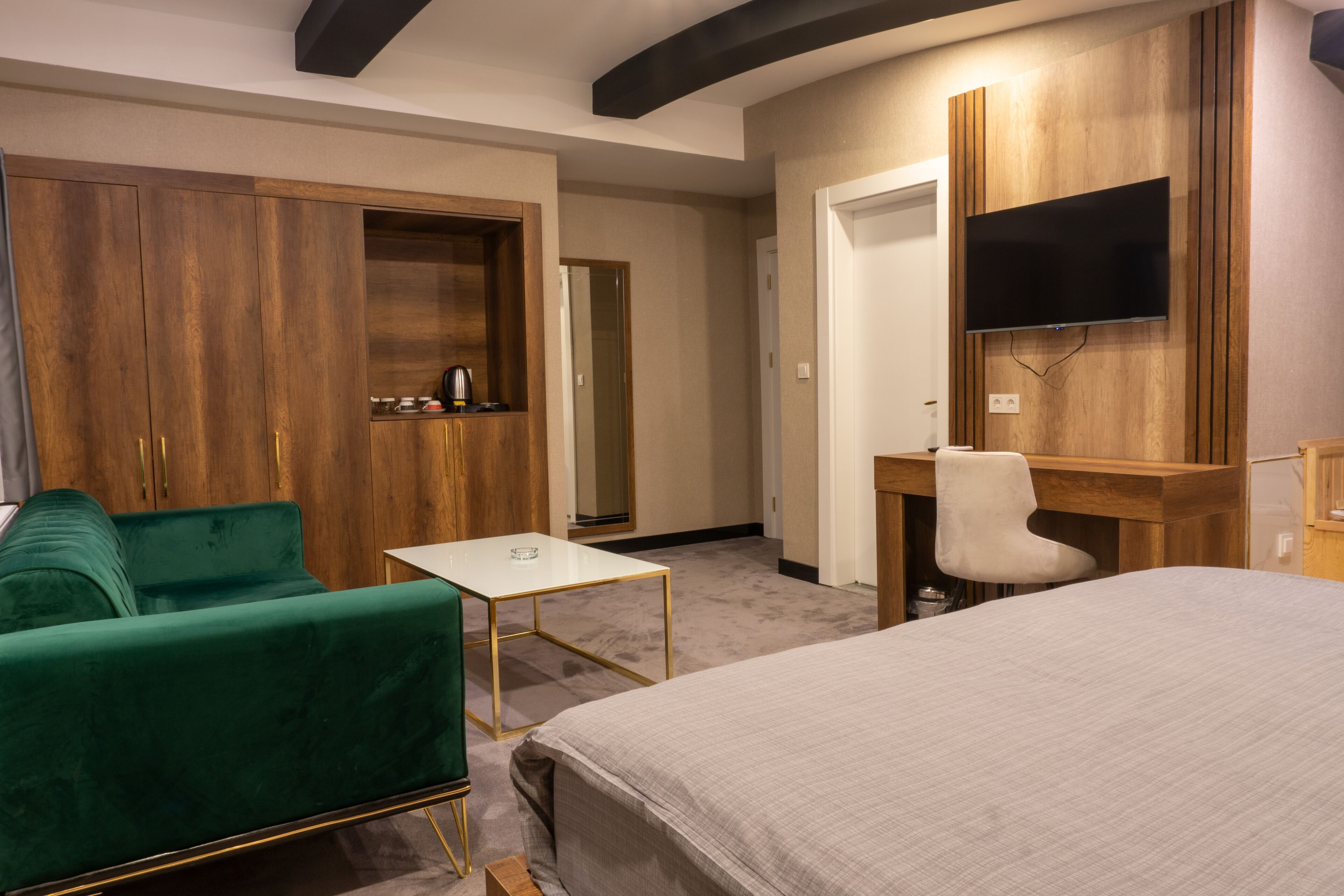 A hotel room with a bed, a desk, a chair, a green sofa, a wooden wardrobe, and a television mounted on the wall.