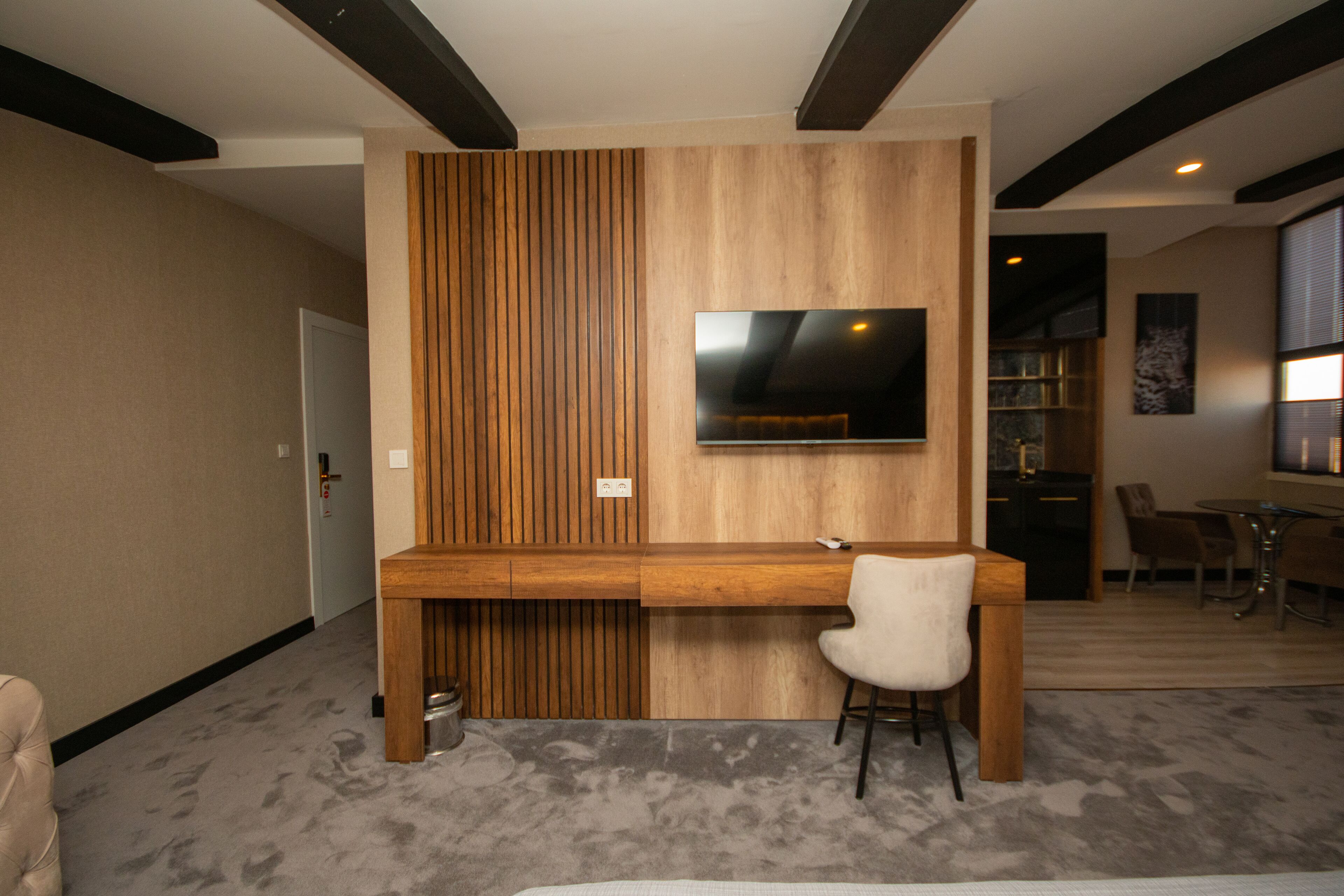 A modern room with a wooden desk, a chair, a television mounted on the wall, and a dining area with chairs and a table.