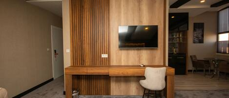 A modern room with a wooden desk, a chair, a television mounted on the wall, and a dining area with chairs and a table.