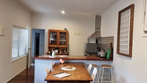 A compact kitchen with a wooden table, bar stools, a cabinet with glass doors, a microwave, and a 'hola' sign on the wall. - Sunset Beach Shack on Surf (Inverloch)