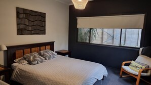 A bedroom with a bed, a nightstand, a chair, a window with a view, and a wall art piece. - Sunset Beach Shack on Surf (Inverloch)