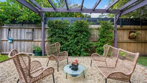 A covered patio with wicker furniture and a small table, surrounded by a wooden fence and greenery. - Sunset Beach Shack on Surf (Inverloch)