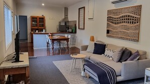 A living room with a sofa, coffee table, and TV. There is a kitchen in the background with a dining table and chairs. - Sunset Beach Shack on Surf (Inverloch)