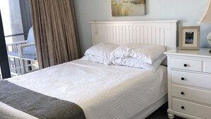 Ein ordentlich bezogenes Bett mit weißer Bettwäsche, eine weiße Kommode mit Schubladen, ein Strandbild und ein Fenster mit Vorhängen.