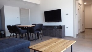 Een moderne woonkamer met een houten salontafel, een bank, een eethoek met tafel en stoelen, en een flatscreen televisie die aan de muur is bevestigd.