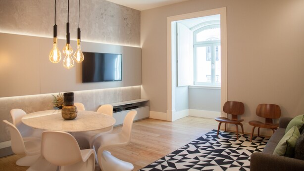 A modern living room with a round dining table, white chairs, a sofa, and a TV mounted on the wall.
