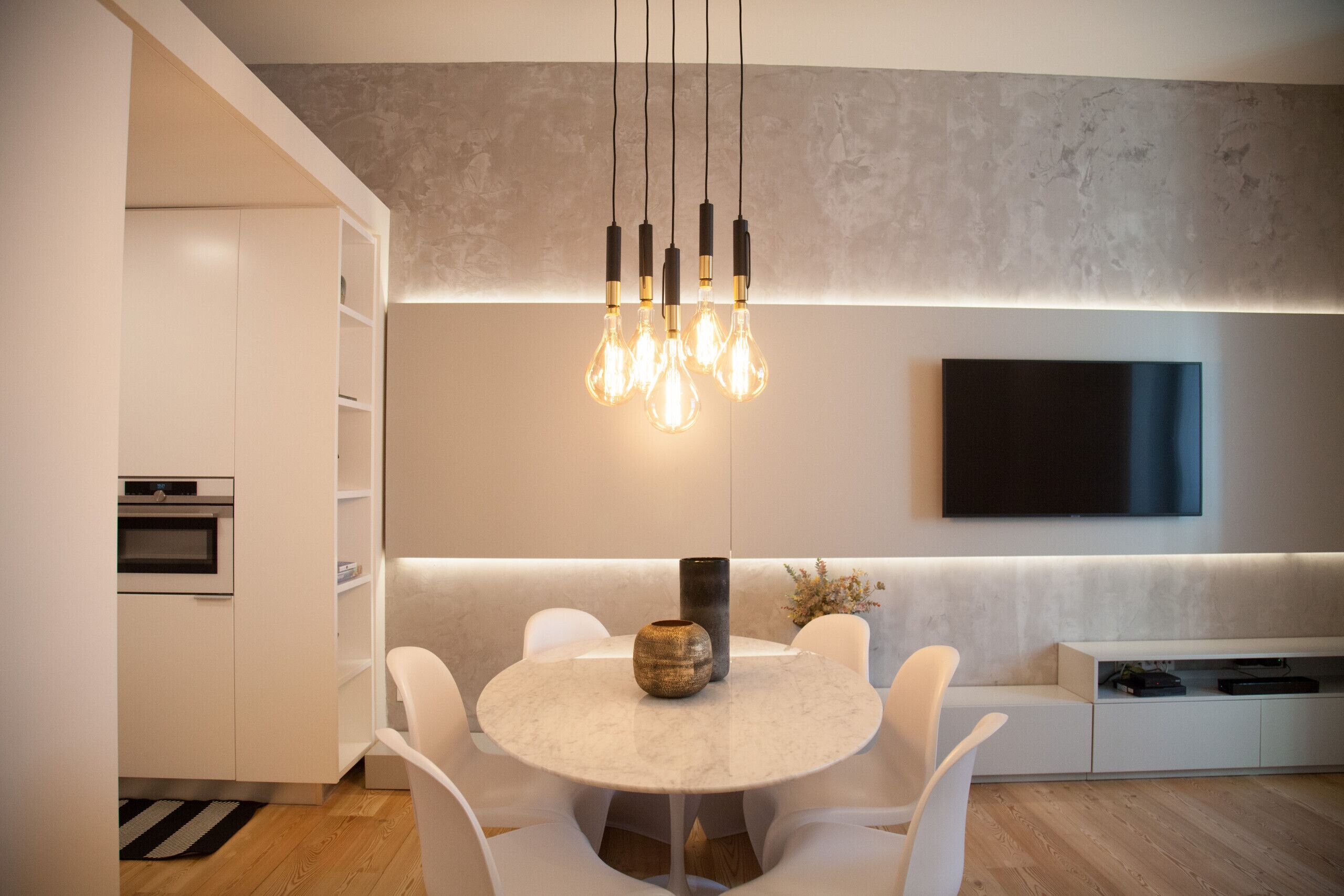 Una cocina moderna con una mesa de comedor redonda, sillas blancas, horno empotrado y un televisor de pantalla plana montado en la pared.