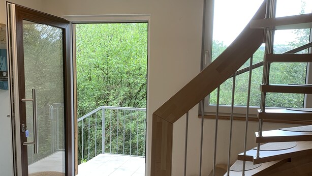 Un interior moderno con un piso de baldosas, una escalera de madera y un vidrio que da a un balcón.