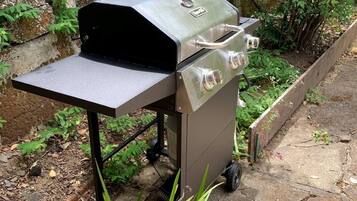 Una parrilla portátil con tapa y quemadores laterales, ubicada al aire libre sobre una superficie de concreto con plantas a su alrededor.