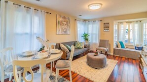 Ein Wohnzimmer mit Esstisch, Stühlen, einer Couch, einem gemusterten Teppich und einem Fenster mit Vorhängen.