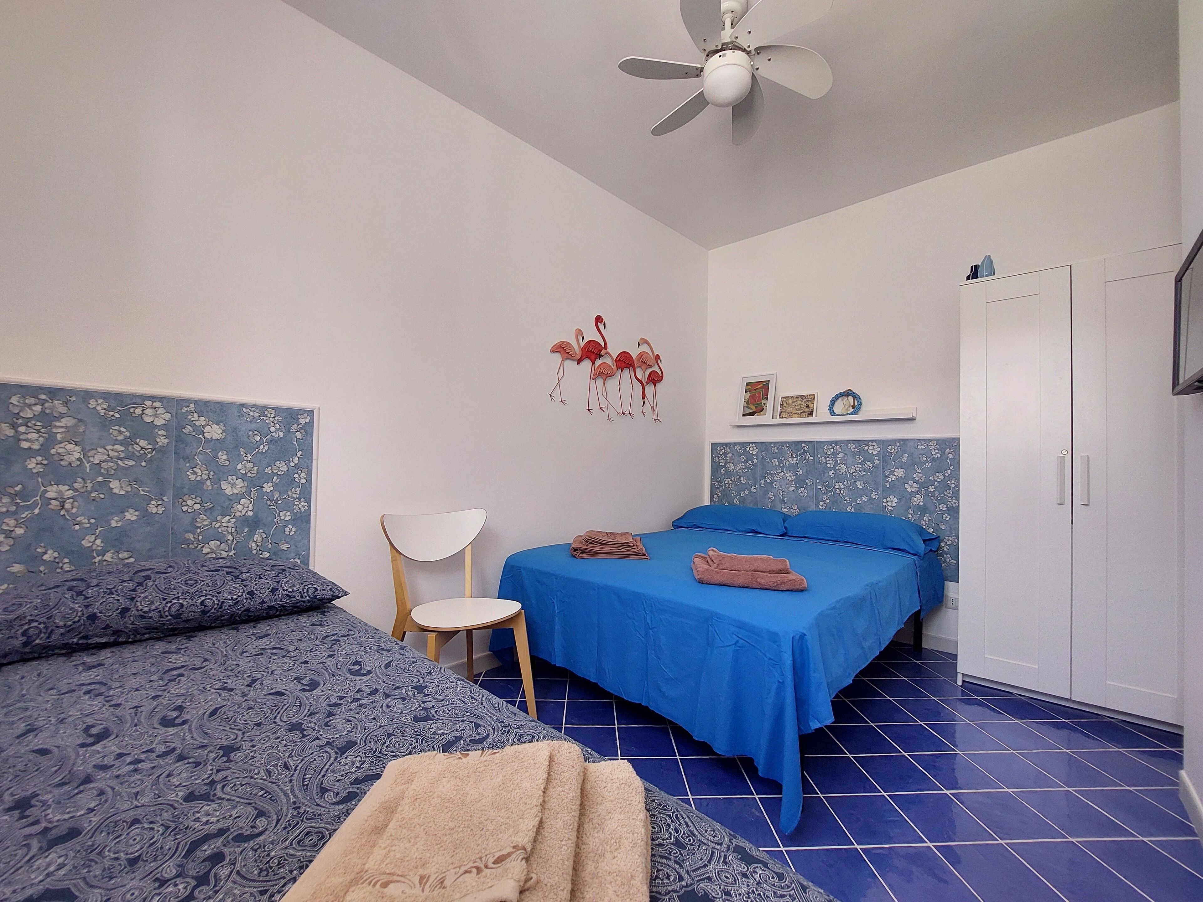Habitación con dos camas, ventilador de techo, una silla, piso de baldosas azules y un armario.