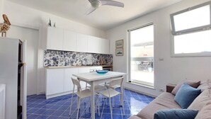 A compact living space with a kitchenette, a dining area with a table and chairs, a sofa with cushions, and a ceiling fan. - AB_Apartment  (Marina di Ragusa)