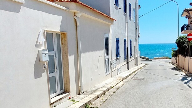 A narrow street with a building featuring a door and windows with shutters, leading to a view of the sea. - AB_Apartment  (Marina di Ragusa)