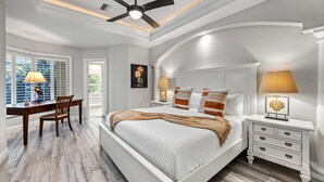 A bedroom with a large bed, a desk, and a chair. There is a ceiling fan, a nightstand with a lamp, and a picture on the wall.