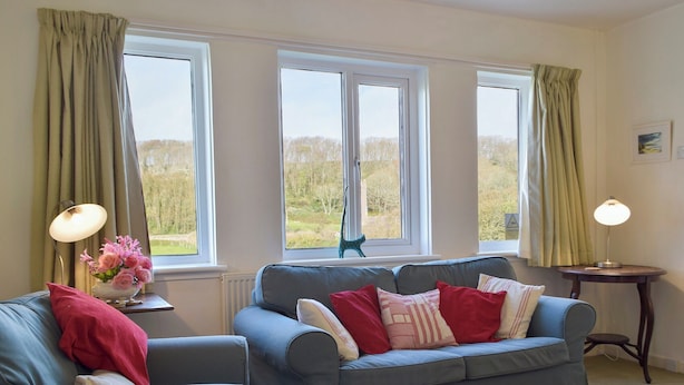 Ein Wohnzimmer mit einem blauen Sofa, einem roten Zierkissen, einem Beistelltisch mit Lampe und Blick ins Freie durch große Fenster.