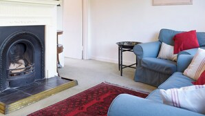 Ein Wohnzimmer mit Kamin, einem blauen Sofa mit roten und gestreiften Kissen, einem kleinen runden Tisch und einem roten Teppich mit Muster.