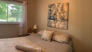 Un dormitorio con cama, una ventana con cortinas, una lámpara y un cuadro abstracto colgado en la pared.