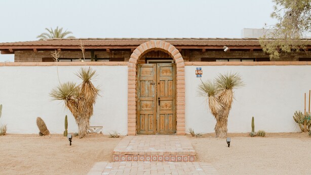 A building with a large wooden door, surrounded by cacti and a paved entrance.